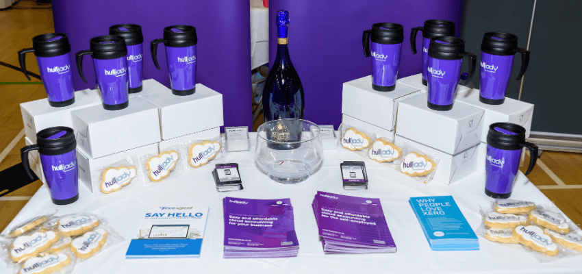 a table topped with purple branded merchandise and a bottle of wine at a business expo